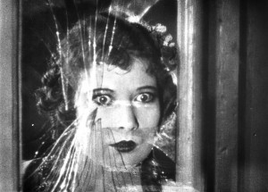 Still from black and white film showing an alarmed woman looking through broken glass